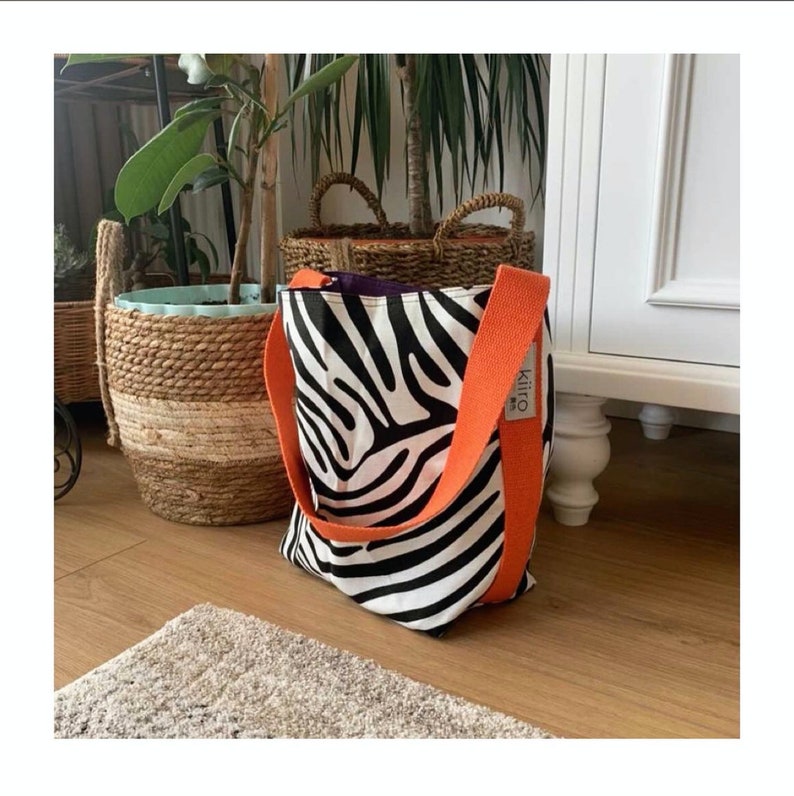 Puede incluir: Una bolsa de tela de color blanco y negro con estampado de cebra y asas naranjas. La bolsa tiene una peque&ntilde;a etiqueta que dice "Kiiro".