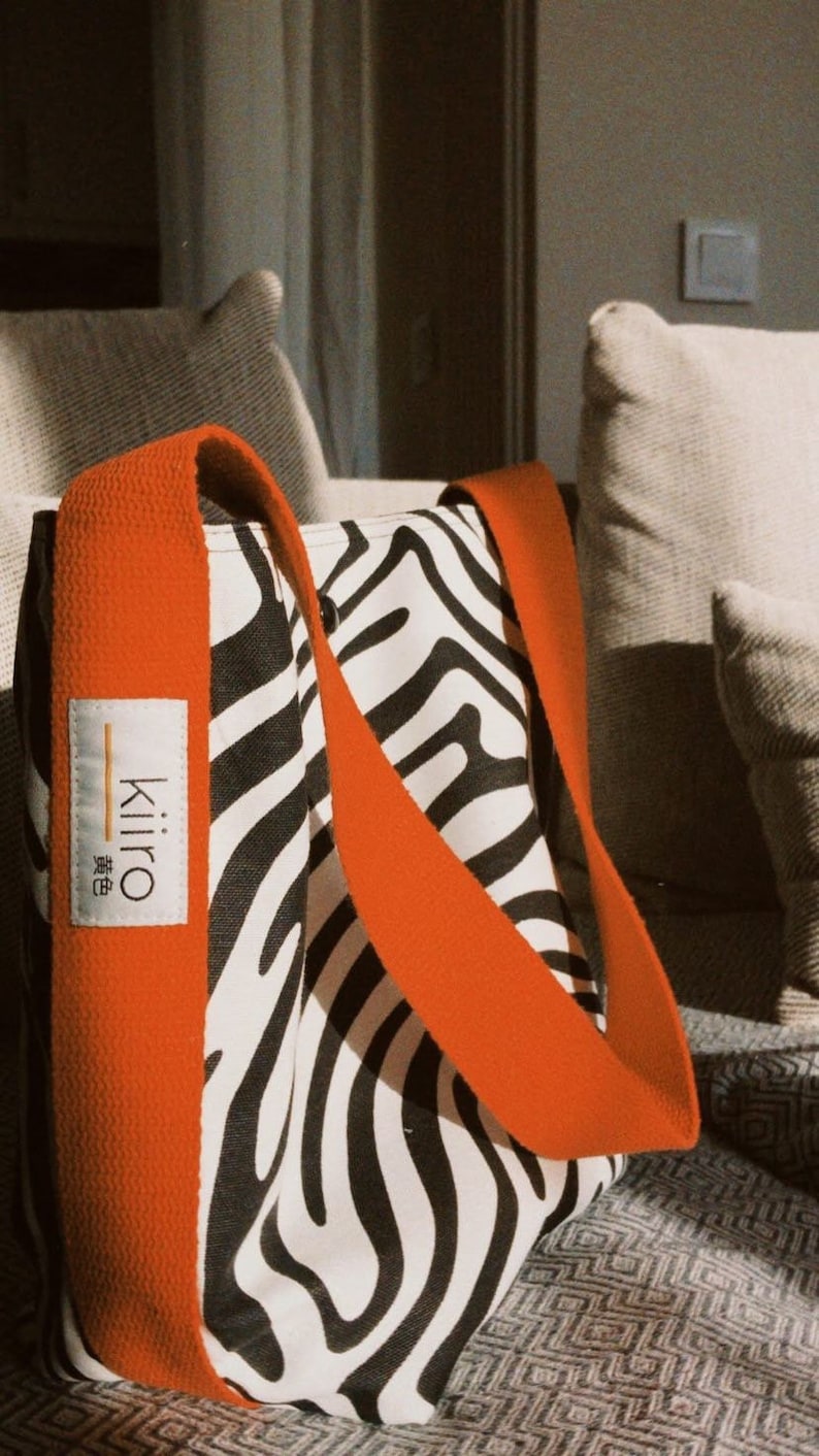 Puede incluir: Una bolsa de mano de lona con estampado de cebra en blanco y negro con correas naranjas. La bolsa tiene una peque&ntilde;a etiqueta blanca con la palabra "Kiiro" y la palabra japonesa para "amarillo" escrita en negro.