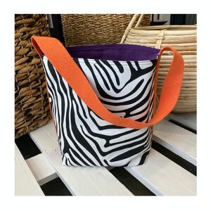 Puede incluir: Una bolsa de mano de lona con estampado de cebra en blanco y negro, con una correa naranja y un forro morado.