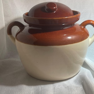May include: A two-handled ceramic crock featuring a glossy brown glazed upper section and lid, contrasted with a smooth cream-colored base. The vessel has a wide opening, a rounded body, and two sturdy side handles for lifting and carrying.
