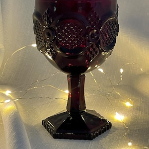 May include: A ruby red glass goblet with ornate embossed detailing. The goblet features a square base, a faceted stem, and a bowl with a textured pattern. The glass is illuminated by warm white fairy lights.