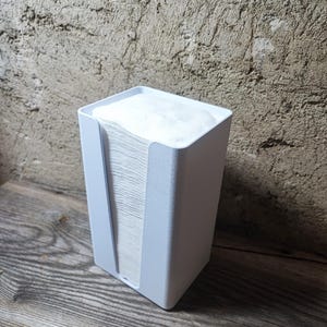 Könnte beinhalten: Ein weißer, rechteckiger Taschentuchspender mit einem vertikalen Schlitz auf der Vorderseite. Die Box ist mit weißen Taschentüchern gefüllt. Der Spender steht auf einer Holzoberfläche, mit einer strukturierten Wand im Hintergrund.