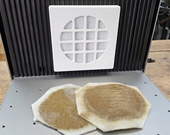 May include: Two round, white, air filters with a hexagonal shape. The filters are dirty and brown with a white border. The filters are sitting on a silver surface.