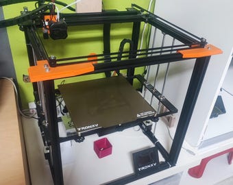 May include: A black 3D printer with orange accents. The printer is on a white surface with a red box and a black display that reads "TRONXY".