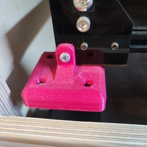 May include: A close-up of a pink 3D printed bracket with four screw holes. The bracket is attached to a black metal frame with screws.