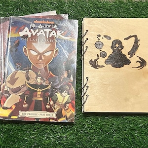 May include: A stack of Avatar: The Last Airbender comic books next to a wooden notebook. The comic books have a colorful cover with the show's characters and the title in English and Chinese. The notebook has a burned design of the Avatar symbol.