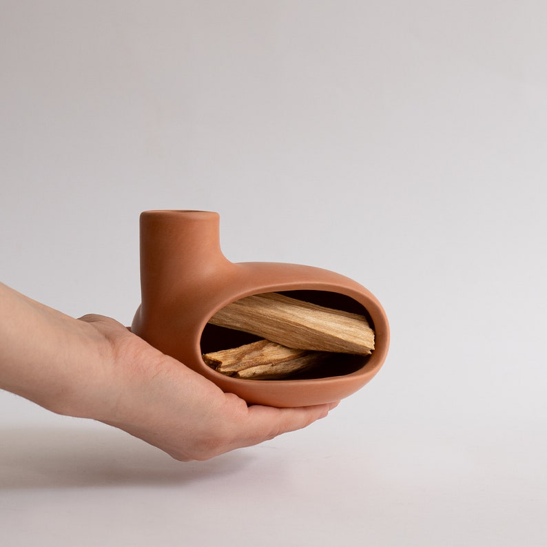 May include: A terracotta ceramic incense holder with a rounded shape and a small opening on the top. The holder is filled with several sticks of incense.