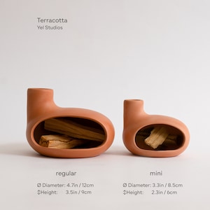 May include: Two terracotta ceramic incense holders, one regular and one mini. The regular holder is 4.7 inches in diameter and 3.5 inches tall. The mini holder is 3.3 inches in diameter and 2.3 inches tall. Both holders have a unique, rounded shape with a hole in the side for incense sticks.