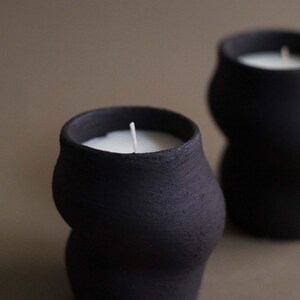 May include: Two black, textured ceramic candles with a unique, curved shape. Each candle has a white wax interior and a single, centered wick. The background is a muted, neutral tone, highlighting the dark candles.