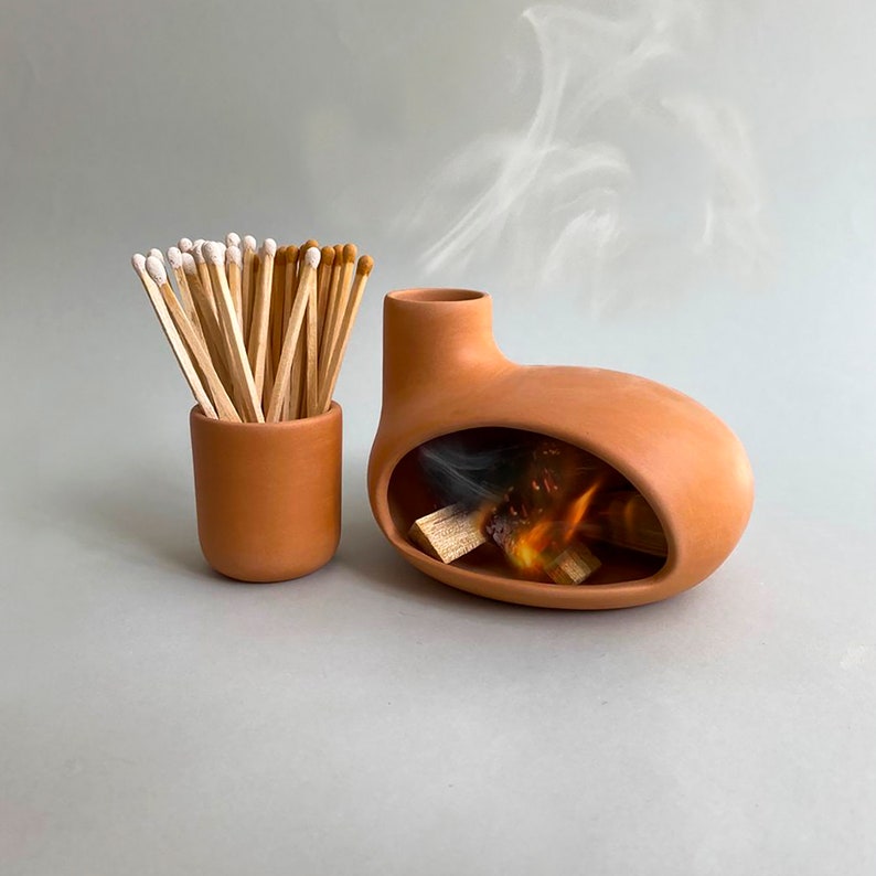 May include: A terracotta fireplace with a small opening and a simulated fire inside. A terracotta jar filled with matches sits next to the fireplace.