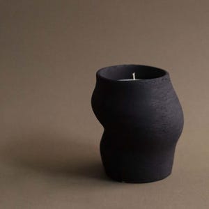 May include: A black, textured candle holder with a unique, curved shape. The candle holder has a wide opening and a small, white candle inside. The background is a solid, muted brown.