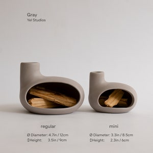 May include: Two gray ceramic vases with a unique design. The larger vase is 4.7 inches in diameter and 3.5 inches tall. The smaller vase is 3.3 inches in diameter and 2.3 inches tall. Both vases have a rounded shape with a small opening at the top and a hollow space in the middle.