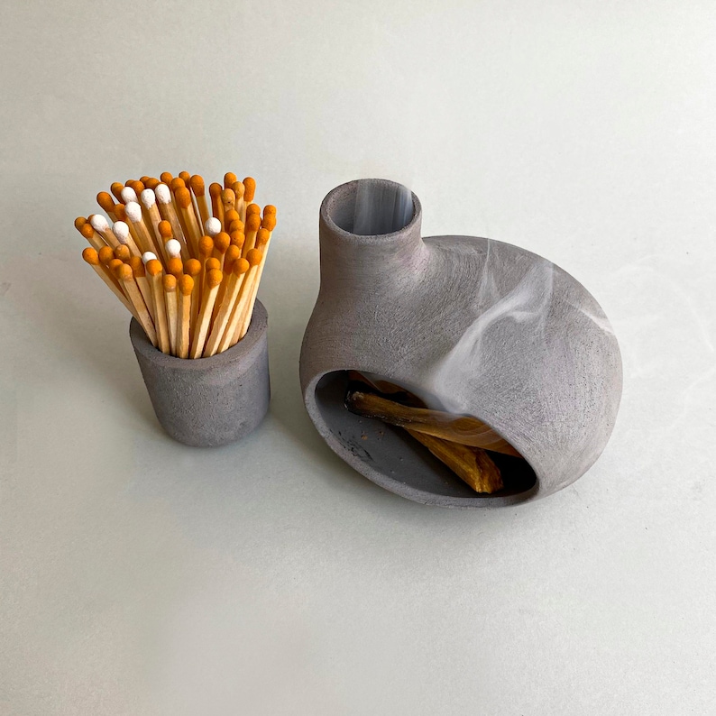 May include: A gray ceramic incense burner with a round opening and a small, gray ceramic match holder filled with wooden matches with orange tips.