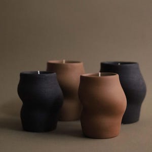 May include: Four sculptural candles in varying shades of brown and black. The candles have a unique, curved shape, resembling a torso. Each candle has a white wick at the top. The background is a solid, muted brown.