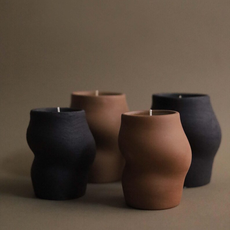 May include: Four sculptural candles in varying shades of brown and black. The candles have a unique, curved shape, resembling a torso. Each candle has a white wick at the top. The background is a solid, muted brown.