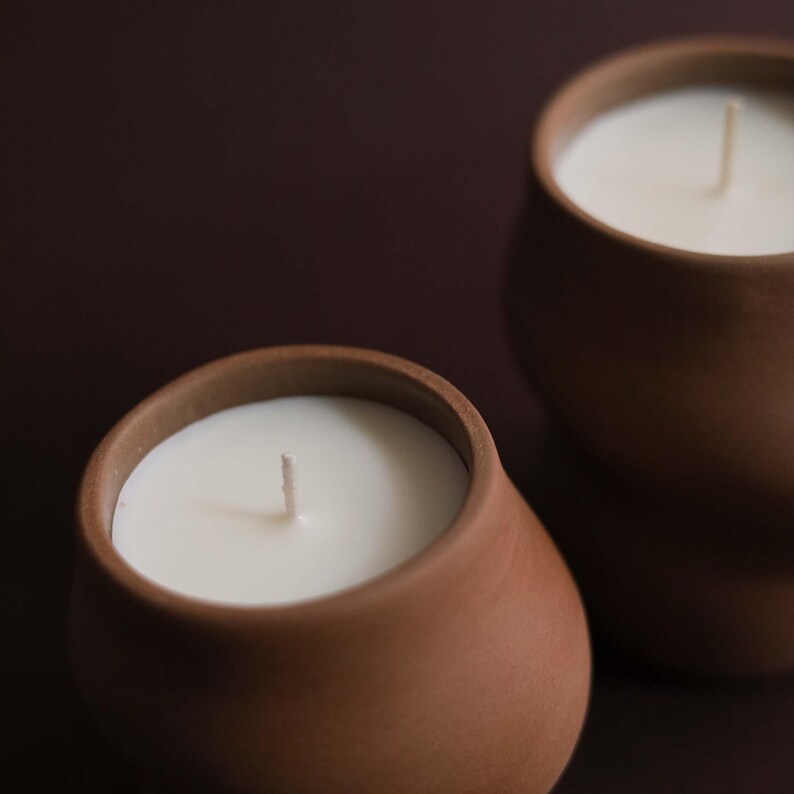 May include: Two brown ceramic candles with white wax and a single wick. The candles have a rounded shape and are set against a dark brown background. The candles are a neutral home decor item.