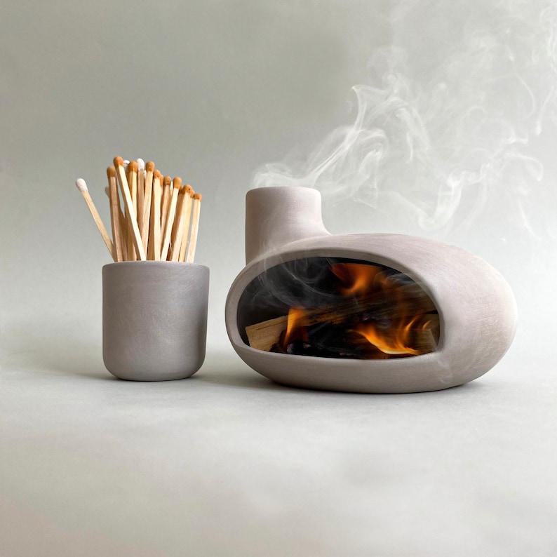 May include: A gray ceramic incense burner with a small opening on the top and a larger opening on the side. The burner is shaped like a small house and has smoke coming out of the top. There is a small gray ceramic match holder next to the burner.