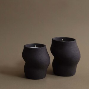 May include: Two black, organically shaped candles with white wicks. The candles have a textured surface and a modern, minimalist design. The candles are different sizes, with one smaller than the other.