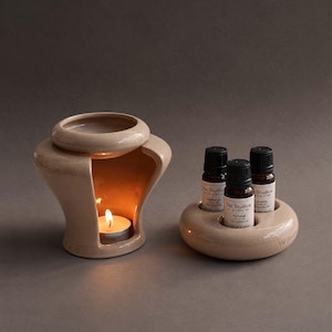 Może przedstawiać: Beżowy ceramiczny kominek do olejków z zapaloną świeczką typu tealight w środku, w towarzystwie pasującej okrągłej ceramicznej podstawki mieszczącej trzy małe buteleczki z ciemnego szkła z olejkami eterycznymi i białymi etykietami.