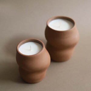 May include: Two terracotta-colored candles with white wax and a single wick. The candles have a unique, rounded shape and are set against a neutral background. The candles are a home decor item.