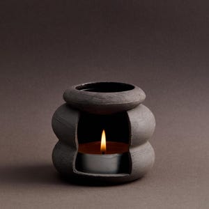May include: A gray ceramic oil diffuser with a small candle burning inside. The diffuser has a rounded, bulbous shape with a wide opening at the top.