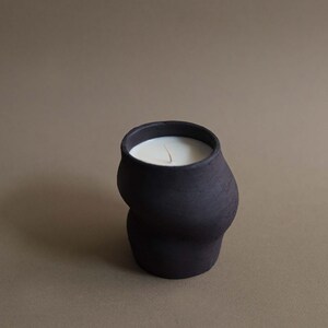 May include: A black ceramic candle with a unique, curved shape. The candle has a white wax interior and a single wick. The dark color of the candle contrasts with the light-colored wax, creating a visually appealing aesthetic. The candle is a decorative item.