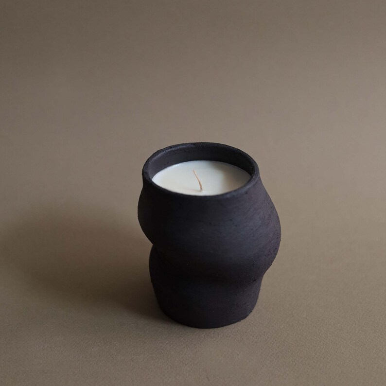 May include: A black ceramic candle with a unique, curved shape. The candle has a white wax interior and a single wick. The dark color of the candle contrasts with the light-colored wax, creating a visually appealing aesthetic. The candle is a decorative item.