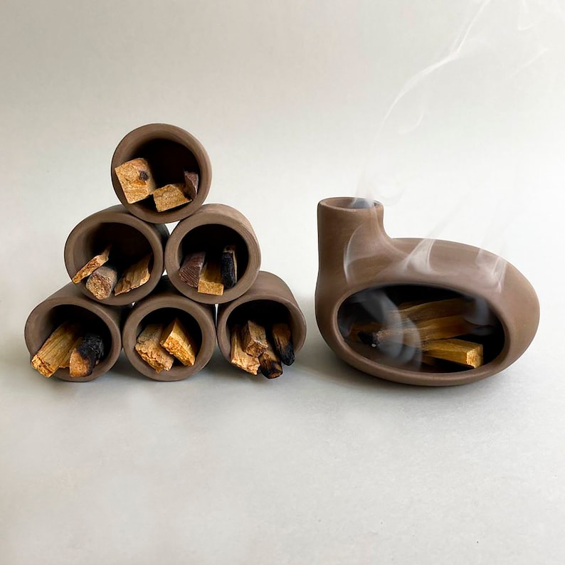 May include: A set of six brown ceramic incense holders arranged in a pyramid shape, each filled with sticks of palo santo wood. A larger brown ceramic incense burner with a rounded shape and a hole in the top is also shown, with smoke rising from the hole.