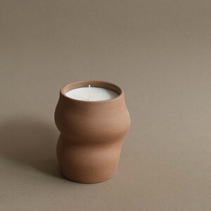 May include: A terracotta-colored candle in a unique, curvy ceramic container. The candle has a white wax surface and a single, centered wick. The container has a smooth, matte finish and a wide opening.