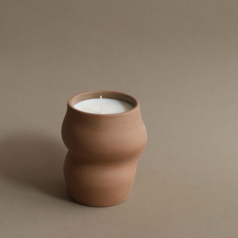 May include: A terracotta-colored candle in a unique, curvy ceramic container. The candle has a white wax surface and a single, centered wick. The container has a smooth, matte finish and a wide opening.