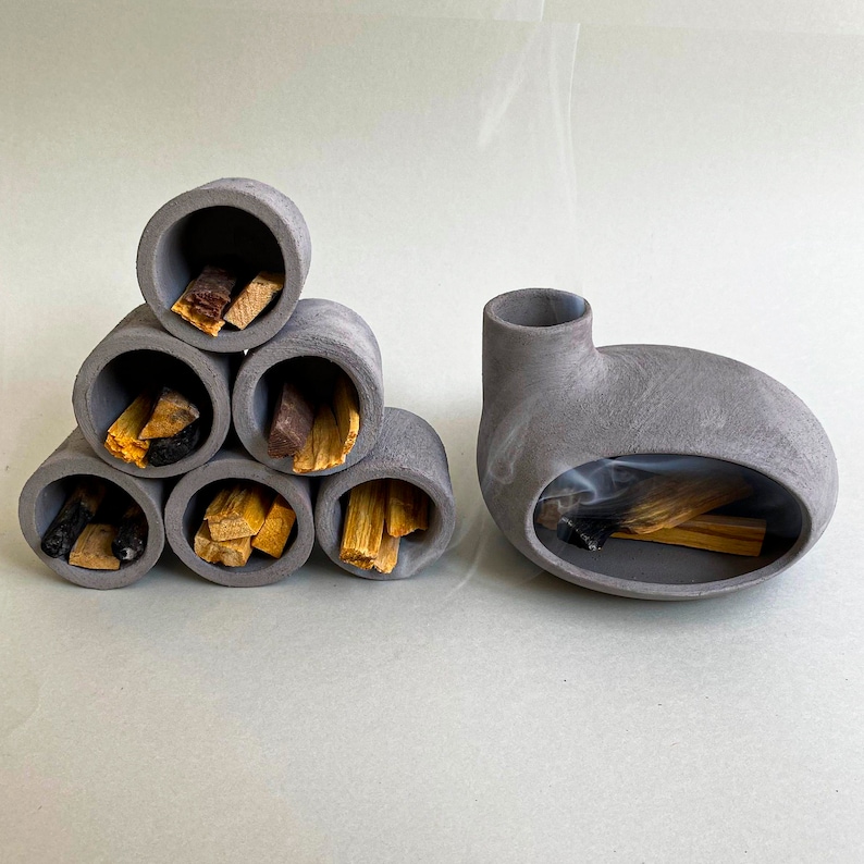 May include: A set of gray ceramic incense holders in the shape of a pyramid and a single, rounded holder. The pyramid holder has five compartments, each filled with sticks of incense. The rounded holder has a single compartment with a stick of incense burning inside.