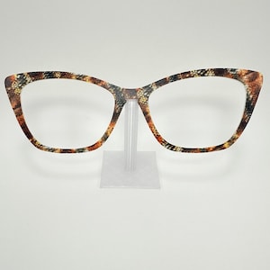 May include: A pair of eyeglasses with a snakeskin pattern in shades of brown, orange, and black. The frames have a cat-eye shape and are displayed on a clear plastic stand. The lenses are clear.