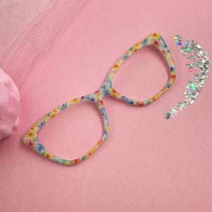 May include: Floral patterned eyeglasses with a light pink background. The glasses have a colorful floral design with blue, yellow, and pink flowers. A small pile of silver glitter is next to the glasses.