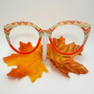 May include: Eyeglasses with a patterned frame in shades of orange, green, and cream, with a red rim. The eyeglasses are displayed on a clear stand, with two orange and yellow artificial leaves in the foreground.
