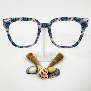 May include: Eyeglasses with a floral pattern in blue and purple, displayed on a clear stand. Below the glasses are several seashells in shades of brown, white, and tan, arranged on a white surface.