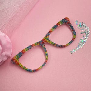 May include: A pair of eyeglasses with a colorful mosaic pattern in rainbow hues. The frames are set against a pink background with a decorative flower and a scattering of iridescent glitter.
