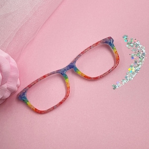May include: Rainbow-colored eyeglasses with a floral pattern. The frames transition through the colors of the rainbow. A small pile of silver glitter is next to the glasses. The background is a solid pink color.