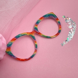 May include: Rainbow-colored eyeglasses with a vibrant, swirled pattern. The frames are set against a pink background with a touch of pink tulle and a scattering of silver glitter.