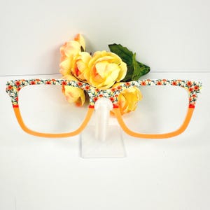 May include: Orange-framed eyeglasses with floral patterned rims. The glasses are displayed with yellow roses and a clear stand. The frames have a bright orange color and are decorated with a white background and red and orange flowers.
