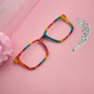 May include: Rainbow-striped eyeglasses with a rectangular frame. The glasses feature a vibrant array of colors, including red, orange, yellow, green, and blue. The background is a soft pink, with a scattering of silver glitter.