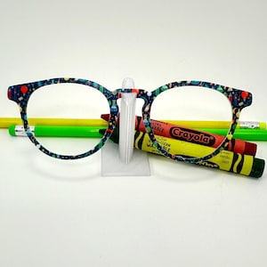 May include: A pair of eyeglasses with a colorful pattern of science-themed elements, resting on a clear stand. Two pencils and three crayons, including a red and yellow Crayola, are in the background. The eyeglasses have a dark blue frame.