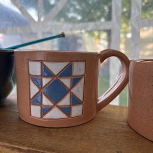 May include: A speckled terracotta-colored ceramic mug featuring a square geometric quilt-inspired pattern on the front. The design consists of blue and white triangles and a central blue diamond shape. The mug has a curved handle on the right side.