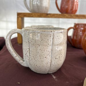 May include: A ceramic mug with a speckled off-white glaze and vertical indentations along the body. The mug features a curved handle and a rounded shape. Additional mugs in off-white and reddish-brown are visible in the background.