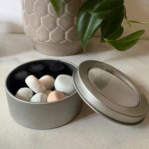 May include: A small, open, silver-colored tin containing several small, light-colored, oval-shaped stones and several small black bottles. The lid of the tin is partially open, revealing a clear window. A decorative pot and green plant are in the background.