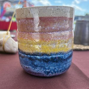 May include: A cylindrical ceramic mug featuring a horizontal gradient of colors, starting with a speckled beige rim, followed by bands of muted red, orange, yellow, and a deep blue base. The surface has a glossy finish with subtle horizontal ridges.
