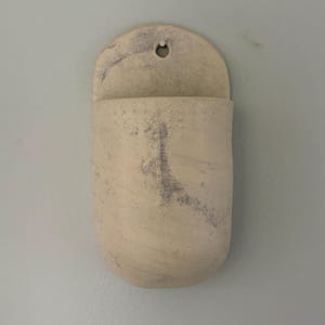 May include: A wall-mounted ceramic pocket planter with a rounded bottom and a flat top edge featuring a small circular hole for hanging. The surface is an off-white, matte finish with subtle, mottled grey and purple-toned mineral-like markings.
