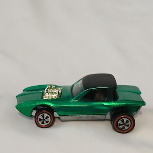 May include: A metallic green toy car featuring a black roof, a silver engine block protruding from the hood, and four wheels with red-line detailing. The vehicle has a low-profile body design with a silver-colored chassis and dark interior.