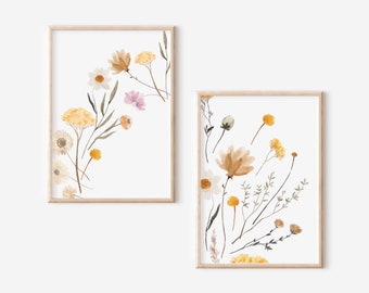 May include: Two framed floral art prints. The prints feature watercolor illustrations of wildflowers in shades of yellow, brown, and pink, with green stems and leaves, set against a white background. The frames are light wood.
