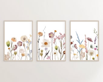 May include: Set of three framed prints featuring watercolor illustrations of wildflowers in shades of pink, yellow, and blue. The prints are arranged in a row on a white wall.