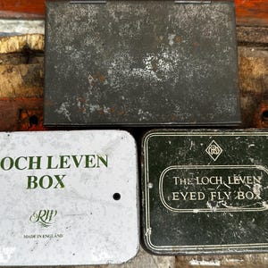 May include: Three rectangular metal storage tins. One is a large, weathered dark grey box. The second is a white tin labelled "LOCH LEVEN BOX" with green text and "MADE IN ENGLAND" printed below. The third is a dark green tin labelled "THE LOCH LEVEN EYED FLY BOX" with white text and a logo.